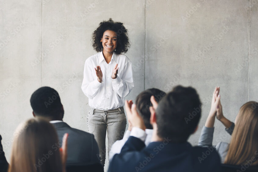 Young female adult standing up clapping in front of a group of adults sitting clapping